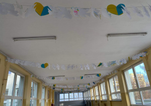 Na zdjęciu widać szkolny korytarz, który ozdobiono pracami uczniów. Wiszą one pod sufitem i są to białe papierowe gołębie, a na nich napisane modlitwy i życzenia.